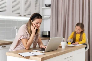 Busy mother using a laptop and phone while her daughter completes homework at the kitchen table, illustrating the need for family support in Dover, Delaware.